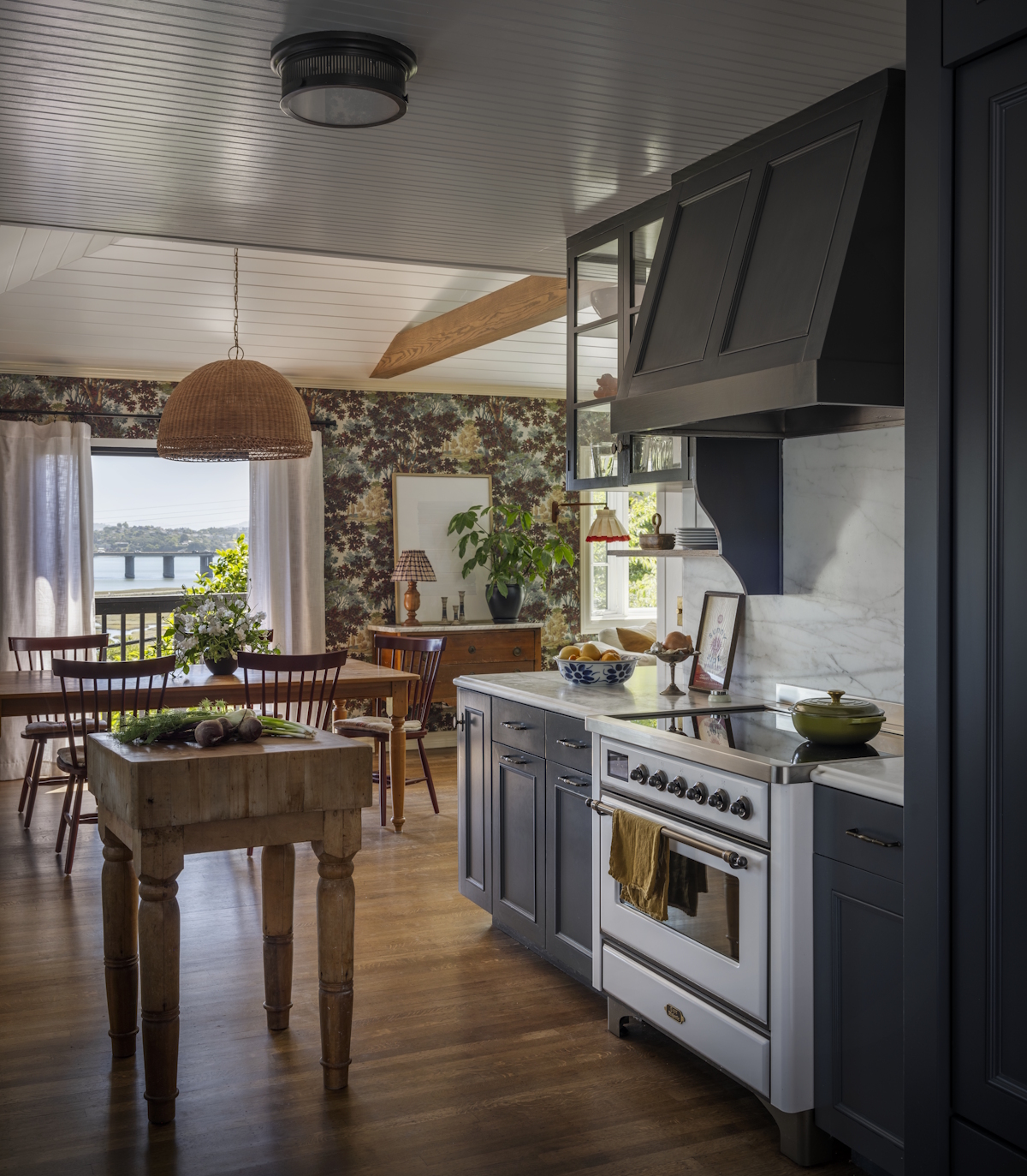 A kitchen featuring gray-blue cabinets and oven hood, a wooden island, and a table with an open layout to the dining and living areas