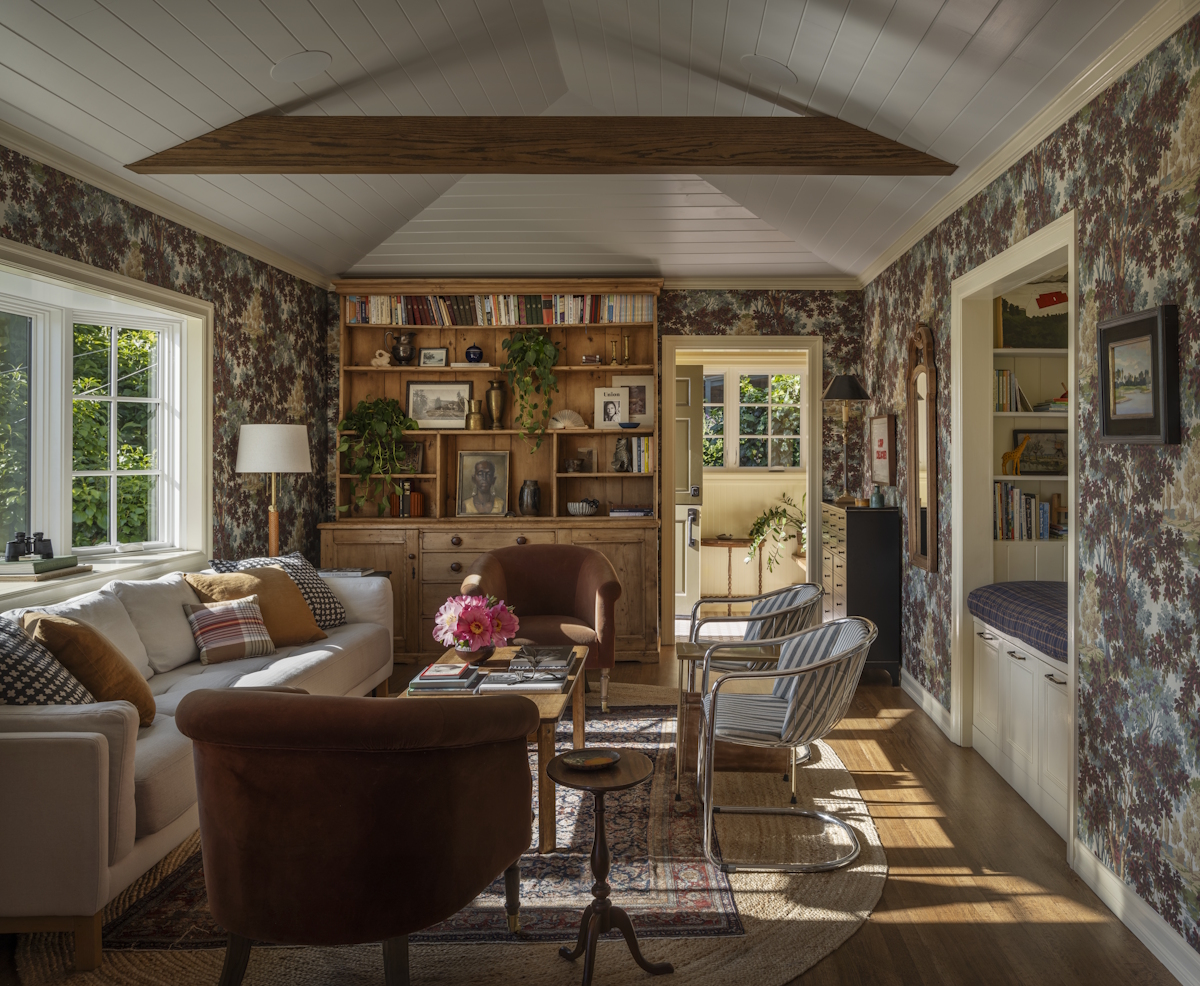 A living room with patterned wallpaper, a wooden wall unit, a light couch with additional chairs around a coffee table, and a built-in reading nook