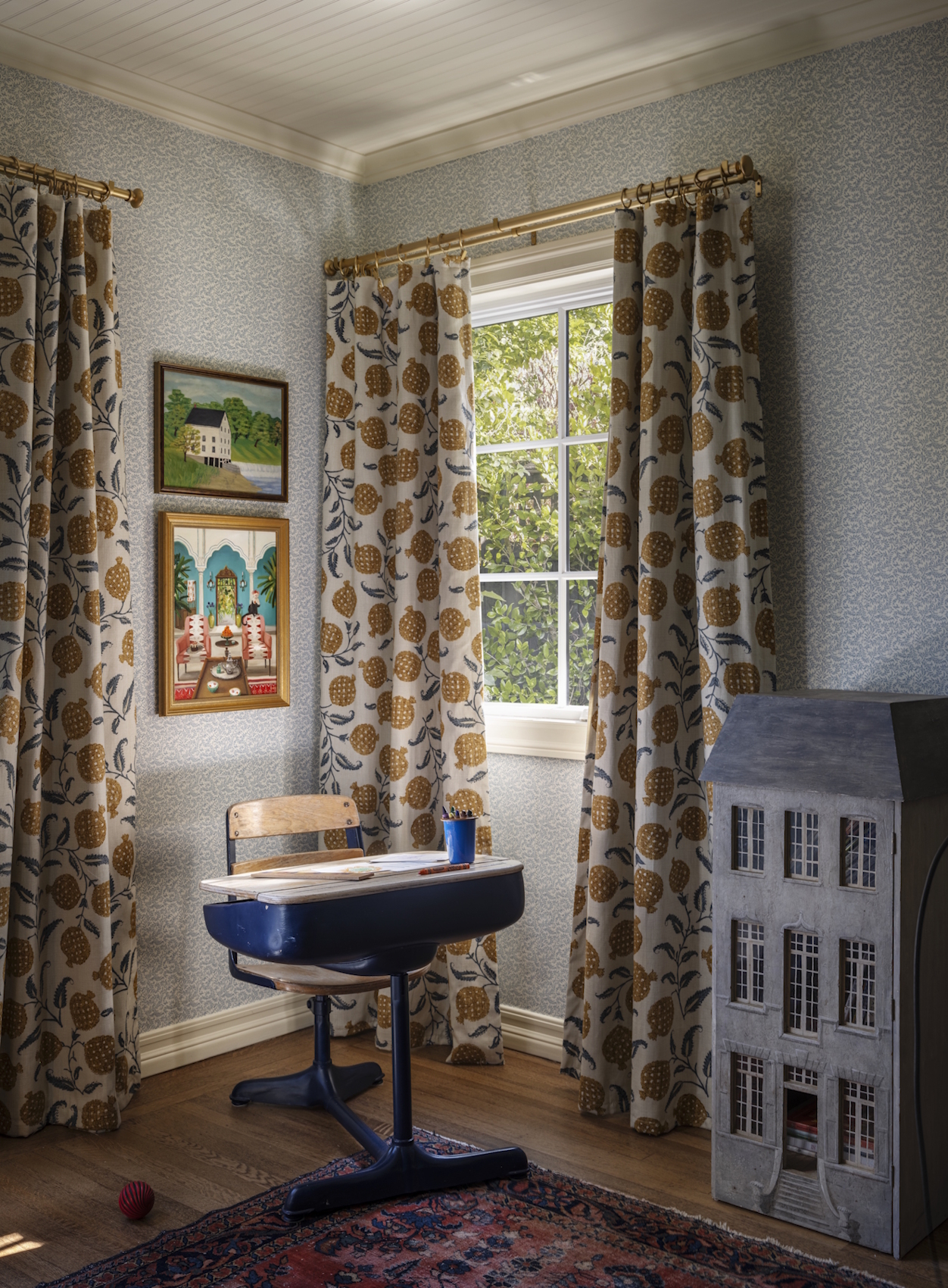 A desk and dollhouse in the corner of a room featuring a window with patterned curtains and wallpaper